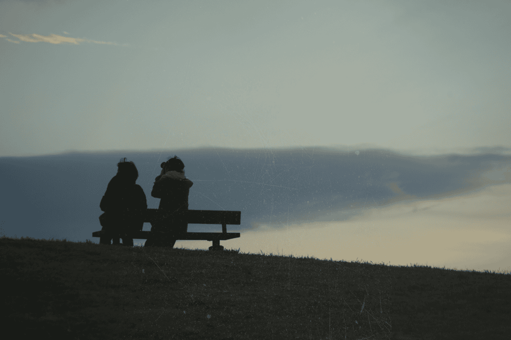 Two people sitting together on a hilltop bench, silhouetted against a dusky sky