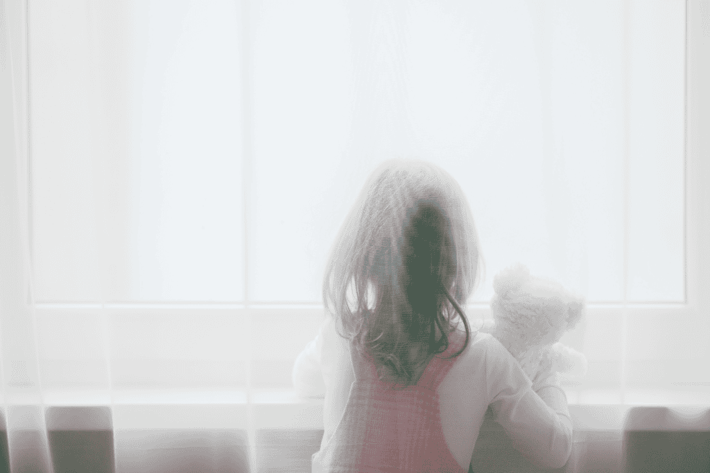 A young girl seen from behind, holding a teddy bear and gazing out a bright window