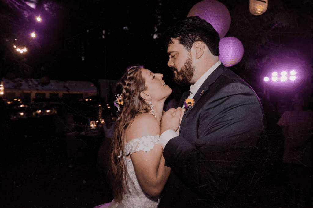 A bride and groom embracing closely, the bride with flowers in her hair, warm glowing lights behind them