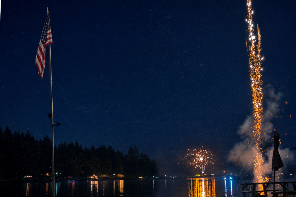 An American flag on a pole beside a lake at night, with fireworks bursting over the water and a picnic table in the foreground